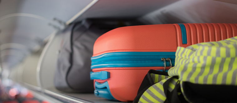 A coral hard-shell cabin bag wedged into an aircraft overhead bin beside a soft grey holdall
